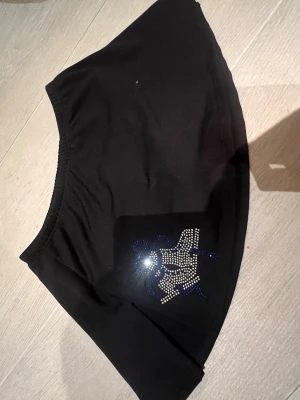 Svart konståkningskjol - Svart kortkjol med strasstenar på ena sidan. Kjolen har en enkel, lätt utsvängd form och är perfekt för dig som gillar en stilren look med en touch av bling.