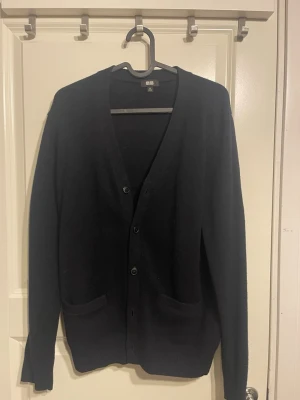 Mörkblå kofta från Uniqlo - Stilren mörkblå kofta från Uniqlo med v-ringning och knäppning framtill. Koftan har två fickor framtill och är tillverkad i ett mjukt, tunt material. Perfekt för lager-på-lager och enkel att matcha med olika outfits. Passar upp till S.