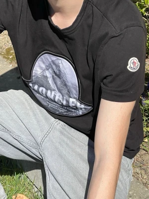 Svart Moncler t-shirt med tryck - Svart t-shirt från Moncler med stort bergstryck och logga på bröstet. Klassisk rund hals och korta ärmar. Moncler-märke på ärmen. Skön och stilren modell som passar perfekt till jeans eller shorts.