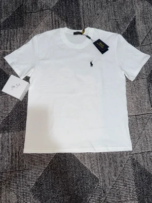 Vit t-shirt från Polo Ralph Lauren - Klassisk vit t-shirt från Polo Ralph Lauren med broderad logga på bröstet. T-shirten har rund halsringning och korta ärmar. Perfekt basplagg med stilren design och mjukt bomullsmaterial.