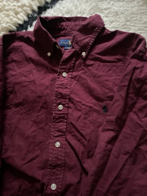 Vinröd skjorta från Ralph Lauren - Säljer en vinröd skjorta från Ralph Lauren med klassisk button-down krage och broderad logga på bröstet. Skjortan har långa ärmar, vita knappar och är tillverkad i mjuk bomull. Perfekt för dig som gillar stilrena plagg med en tidlös touch.