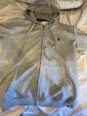 Grå zip hoodie från Polo Ralph Lauren - Snygg grå hoodie med dragkedja från Polo Ralph Lauren. Klassisk design med huva, dragsnören och kängurufickor. Liten mörkblå logga broderad på bröstet. Tillverkad i mjukt bomullsmaterial som är perfekt för chill dagar. Den har använts 2 gånger så det är riktigt bra skick. Storleken är M, pris kan diskuteras. Vid snabbt köp kan det bli billigare.