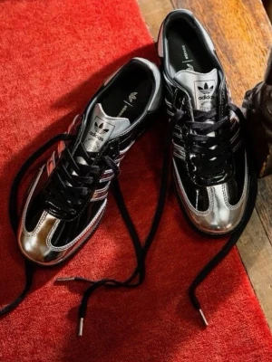 Adidas x Wales Bonner svarta sneakers - Snygga sneakers från Adidas x Wales Bonner med svart och silverfärgad ovandel i glansigt material. Klassiska tre ränder i silver på sidan, svarta skosnören och låg profil. Perfekta för dig som vill sticka ut med en unik och trendig look.