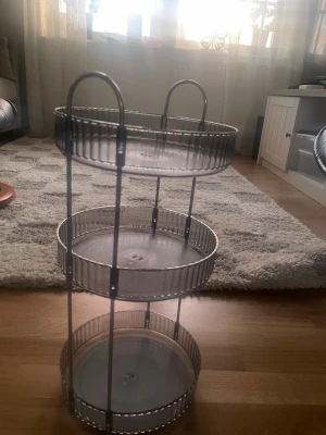 Trestegs rund hylla i metall - Säljer en snygg trestegs rund hylla i silverfärgad metall. Hyllan har tre cirkelformade plan med ribbade kanter och två handtag upptill. Perfekt för förvaring av småprylar eller dekorationer i hemmet.