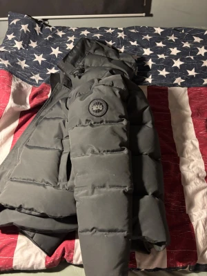 Mörkgrå dunjacka från Canada Goose - Snygg mörkgrå dunjacka från Canada Goose med klassisk patch på ärmen och hög krage. Jackan har en puffig design och är perfekt för kalla vinterdagar. Tillverkad i slitstarkt material och fylld med dun för extra värme.