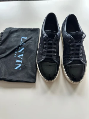 Lanvin marinblå - Snygga svarta sneakers från Lanvin med ovandel i mocka och en glansig, lackad tå. Skorna har vita sulor och klassisk snörning. Kommer med original dustbag från Lanvin. Perfekta för dig som gillar stilrena och exklusiva sneakers.