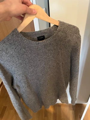 Grå stickad tröja från J.Crew - Säljer en stilren gråmelerad stickad tröja från J.Crew. Tröjan har rund halsringning, långa ärmar och är gjord i shetlandsull. En defekten är den lilla skadan där vid ärmen.