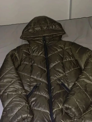 Olivgrön pufferjacka från Amisu - Säljer en olivgrön pufferjacka från Amisu med huva och dragkedja framtill. Jackan har två snedställda fickor med dragkedja och ett quiltat mönster. Materialet är glansigt och ger en cool look till vinterns outfits.