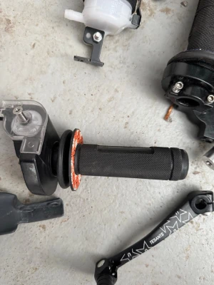 Svart gasreglage  - Ett svart gasreglage med grepp i gummi och plast, samt en svart vevarm från med vitt mönster. Delarna är avsedda för cykel eller moped och har detaljer i metall och plast. Perfekt för dig som gillar att meka med fordon.