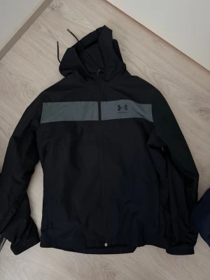 Svart vindjacka från Under Armour - Snygg svart vindjacka från Under Armour med huva och dragkedja framtill. Jackan har en grå panel över bröstet med logga och justerbara snören i huvan. Perfekt för blåsiga dagar och sportiga outfits.