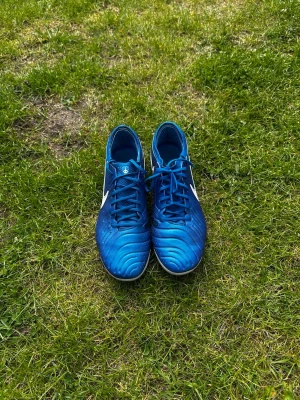 Blå Nike Tiempo fotbollsskor - Säljer ett par blå Nike Tiempo fotbollsskor med vita detaljer och svart Nike-logga på sidan. Skorna har klassisk snörning och dobbar för spel på gräs. Ovandelen är i syntetmaterial med struktur för bättre bollkontroll. Perfekta för dig som vill ha stil och funktion på planen.