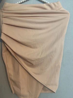 Beige omlottkjol med drapering - Trendig beige kortkjol med snygg omlott-effekt och draperade detaljer framtill. Kjolen har en asymmetrisk nederkant och är tillverkad i ett mjukt, stretchigt material som ger en bekväm passform. Perfekt för dig som gillar stilrena och moderna plagg.