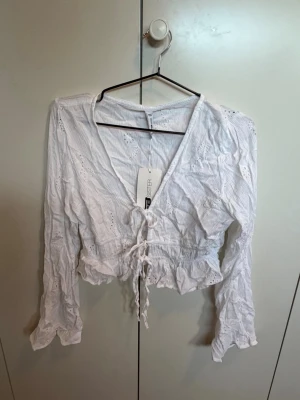 Vit croppad topp från FB Sister - Säljer en vit croppad topp från FB Sister med v-ringning och snörning framtill. Toppen har långa ärmar, broderade detaljer och volangkant nedtill. Perfekt för dig som gillar en romantisk och trendig stil.