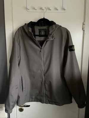 Grå vindjacka från St0ne Island - Snygg grå vindjacka från Stone Island med huva och dragkedja framtill. Jackan har två sidofickor och den klassiska Stone Island-loggan på vänster ärm. Tillverkad i ett lätt och vindtätt material, perfekt för blåsiga dagar.