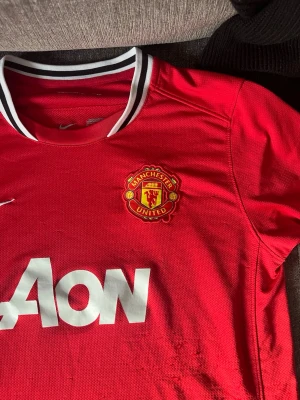 Manchester United matchtröja Nike - Röd Manchester United matchtröja från Nike med klubbmärke på bröstet och vit AON-logga. Tröjan har svartvit krage och är tillverkad i Dri-FIT-material som andas. Perfekt för fotbollsträning eller att bära på matchdag.
