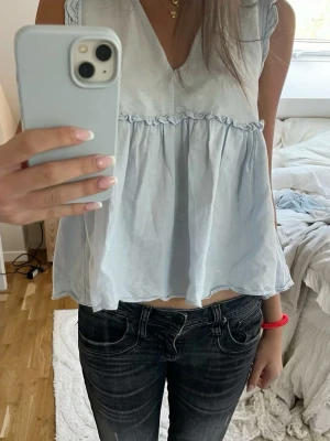 Ljusblå volangblus med v-ringning - Supersöt ljusblå blus med v-ringning och volangdetaljer. Blusen är ärmlös och har en luftig, flowy passform som känns perfekt för varmare dagar. Passar till både jeans och kjol för en chill look.