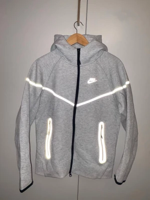 Grå Nike hoodie med reflexdetaljer - Snygg grå hoodie från Nike med dragkedja, huva och reflexdetaljer på bröst, rygg och fickor. Perfekt för dig som gillar sportig stil och vill synas i mörkret. Mjukt material och två fickor med dragkedja framtill.