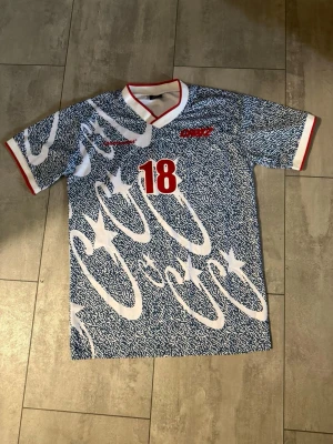 Cortiez fotboll Jersey - Säljer en blåvit fotbollströja med unikt mönster och röda detaljer. Tröjan har nummer 18 på bröstet, röd och vit krage samt röda kantband på ärmarna. Perfekt för dig som gillar sportiga och färgstarka plagg. Size är XL men passar mest som L eller också M