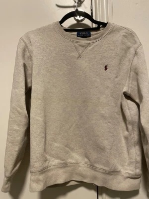 Grå sweatshirt från Polo Ralph Lauren - Klassisk grå sweatshirt från Polo Ralph Lauren med rund halsringning och broderad logga i vinrött på bröstet. Tröjan har långa ärmar och ribbade muddar vid ärmslut och nederkant. Perfekt för en avslappnad och stilren look.