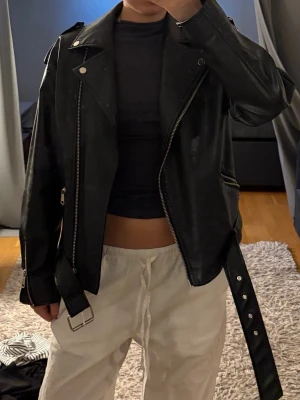 Svart biker skinnjacka med bälte - Snygg svart skinnjacka i klassisk bikerstil med breda slag, silverfärgade dragkedjor och detaljer samt ett justerbart bälte i midjan. Jackan har axelklaffar och flera fickor med dragkedja.