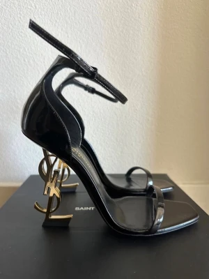 YSL HEELS - Säljer ett par svarta pumps från Saint Laurent med öppen tå och smala remmar. Klacken är i guld och formad som YSL-loggan vilket ger en unik och lyxig look. Skorna är i blankt läder och har en justerbar ankelrem. Och ja den är 100% äkta! Skickar snabbt 