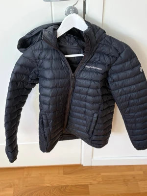 Svart dunjacka Peak Performance - Snygg svart dunjacka från Peak Performance med huva och dragkedja framtill. Jackan har diskret logga på bröstet och ärmarna, quiltad design och är lätt vadderad för extra värme.