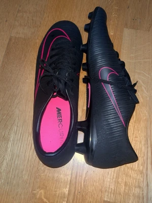 Nike svarta fotbollsskor (nytt skick) - Säljer ett par svarta Nike Mercurial fotbollsskor med rosa detaljer och klassisk Swoosh-logga. Skorna har snörning, dobbar för spel på gräs och en snygg, modern design. Insidan är rosa och ovandelen är i syntetmaterial med struktur för bättre bollkontroll. Storlek 45 passar 44