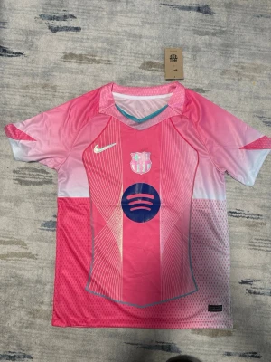 FC Barcelona rosa matchtröja Nike - Snygg FC Barcelona matchtröja i rosa och vitt med tunna linjer och blå detaljer. Tröjan har klubbmärke, Nike-logga och Spotify-logga på bröstet. Tillverkad i lätt och ventilerande polyester, perfekt för fotboll eller träning.