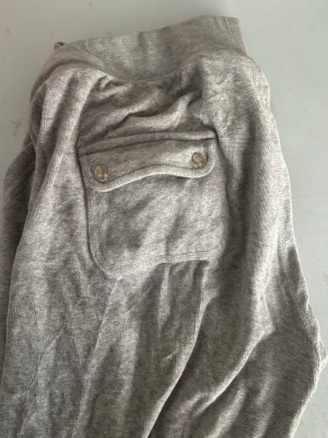 Grå mjukisbyxor med fickor - Säljer ett par grå mjukisbyxor i mjukt bomullsmaterial. Byxorna har dragsko i midjan, två sidofickor och en bakficka med lock och knappar. Perfekta för chill dagar eller när du vill vara bekväm.