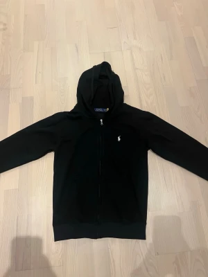 Svart hoodie från Polo Ralph Lauren - Svart hoodie med dragkedja från Polo Ralph Lauren. Klassisk design med huva och vit broderad logga på bröstet. Tillverkad i mjukt material som känns skönt mot huden. Perfekt för en avslappnad och stilren look.