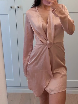 Ljusrosa omlottklänning i satin - Supersnygg ljusrosa omlottklänning från PrettyLittleThing i glansig satin. Klänningen har lång ärm, v-ringning och kort längd med draperad detalj framtill. Perfekt för dig som vill ha en chic och trendig look.