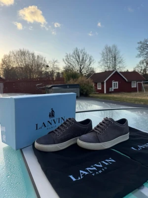 Gråa Lanvin sneakers med vit sula - Stilrena grå sneakers från Lanvin med klassisk låg profil och vit sula. Skorna har ovandel i mocka och textil, samt matchande grå skosnören. Perfekta för dig som gillar en clean och exklusiv look. Kommer med originalkartong och dustbags.
