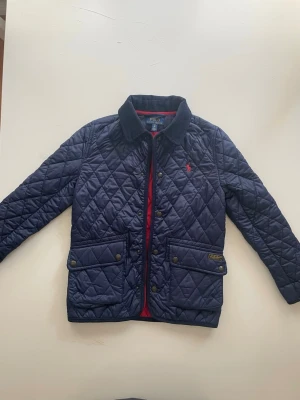 Polo Ralph Lauren jacka - Quiltad mörkblå vindjacka från Polo Ralph Lauren med röd insida och röd logga på bröstet. Jackan har två stora fickor med knappar, ståkrage i manchester och snygga detaljer. Perfekt för en cool och stilren look. Storleken passar 8-10 åringar 