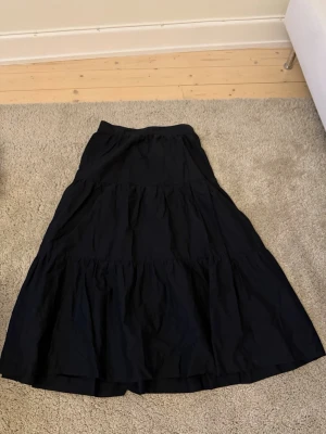 Svart långkjol från H&M Divided - Säljer en svart långkjol från H&M Divided med volangdetaljer och resår i midjan. Kjolen är tillverkad i ett lätt och luftigt bomullstyg, perfekt för en avslappnad och trendig look.