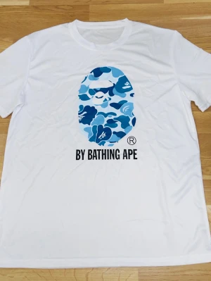 Vit BAPE t-shirt med blått camomönster - Säljer en vit t-shirt från BAPE med det klassiska apansiktet i blått camomönster på bröstet och texten 'BY BATHING APE' under. T-shirten har rund hals och korta ärmar. Perfekt för dig som gillar streetwear och vill sticka ut.