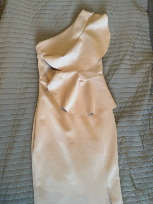 Champagnefärgad oneshoulder klänning - Säljer en elegant champagnefärgad klänning med oneshoulder och volangdetaljer. Klänningen har en slits framtill och är figurnära med snygg passform. Perfekt för dig som vill sticka ut med en unik och stilren look.