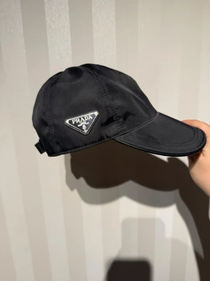 Svart keps från Prada - Snygg svart keps från Prada med klassisk böjd skärm och justerbar rem baktill. Kepsen är tillverkad i nylon och har Pradas ikoniska metallogo på sidan. Perfekt för dig som vill ha en stilren accessoar med lyxig känsla.