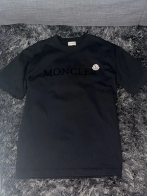 Svart Moncler t-shirt med logga - Svart t-shirt från Moncler med stor svart logotyp på bröstet och klassisk Moncler-patch på vänster sida. T-shirten har rund hals och korta ärmar, tillverkad i mjuk bomull för en clean och stilren look.