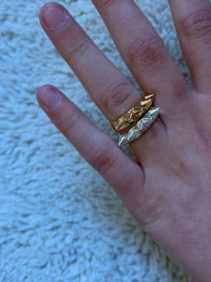 Två ringar med nitar i guld och silver - Säljer två coola ringar med nitar, en i guld och en i silverfärg. Båda ringarna har en edgy design med spetsiga detaljer runt om. Perfekta för att ge din stil en tuff touch. Materialet är metall.