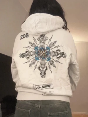 Vit hoodie med grafiskt tryck - Säljer en vit hoodie från Li-Ning med stor grafisk design i svart och blått på ryggen och mindre tryck på bröstet. Hoodien har huva med dragsko och en stor magficka. Perfekt för dig som gillar streetwear och unika prints.
