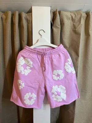 Denim Tears cotton Wreath Shorts  - Säljer nu ett par Denim Tears Mono Cotton Wreath Shorts i färgen rosa. Shortsen är från FW24-kollektionen och har det ikoniska "Cotton Wreath"-trycket i vitt. Detaljer: • Modell: Mono Cotton Wreath Short • Färg: Pink / Rosa med vitt tryck • Storlek: Medium (M) • Skick: Toppskick / Nyskick. Endast testade men säljes då de tyvärr inte passade mig. • Material: Kraftig och högkvalitativ bomull (French Terry-insida). • Tillverkade i: Portugal.