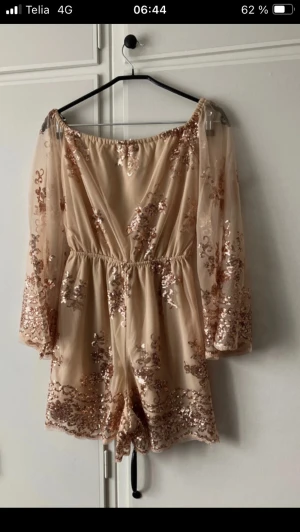 Beige jumpsuit med paljetter - Superfin beige jumpsuit med vida ärmar och glittriga beigeguldfärgade paljetter. Off shoulder-modell med resår i midjan och transparenta ärmar. Perfekt för dig som vill sticka ut med en trendig look! Supervacker! Slutsåld.