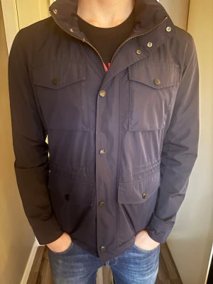 Mörkblå field jacket Bondelid Gerrard - Stilren mörkblå vindjacka från Bondelid med fyra framfickor och dragkedja samt tryckknappar. Jackan har hög krage och är tillverkad i ett lätt, vattenavvisande material. Perfekt för blåsiga dagar och enkel att matcha med jeans eller sneakers. Modelnamn: Bondelid Gerrard Field Jacket. Nypris 1800kr. Ställ gärna frågor kring jackan eller pris!