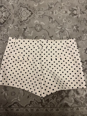 Vita prickiga shorts med dragkedja - Snygga vita shorts med svarta prickar och klassisk passform. Shortsen har bälteshällor, två bakfickor och stängs med dragkedja och knapp framtill. Perfekta för sommardagar och ger en retrovibe till din outfit.