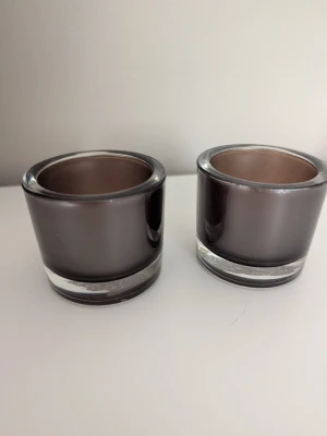 Två rökfärgade ljuslyktor i glas - Säljer två stilrena ljuslyktor i rökfärgat glas. De har en rund form och tjock botten, perfekta för värmeljus. Passar snyggt på bordet eller i fönstret för en mysig känsla.