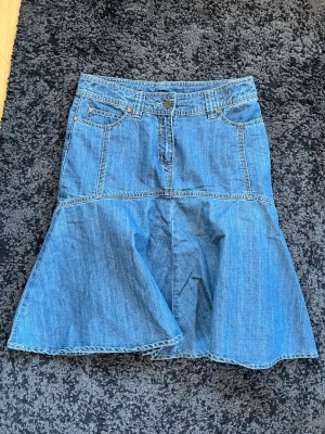 Blå jeanskjol med utsvängd form KappAhl - Snygg blå jeanskjol med utsvängd nederdel och klassiska fickor fram och bak från KappAhl i storlek M. Kjolen har bälteshällor och knappstängning i midjan. Perfekt för dig som gillar en avslappnad men trendig stil. Midja: 39cm. Längd: 58cm.