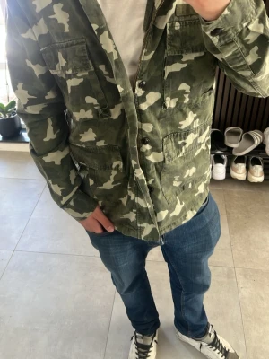 Grön camo overshirt med fickor - Säljer en grön overshirt med camouflagemönster och fyra stora fickor framtill. Skjortjackan har knäppning med knappar och är tillverkad i ett slitstarkt bomullstyg. Perfekt för lager-på-lager och streetwear-stil.