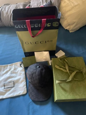 Gucci keps med GG-mönster - Snygg svart keps från Gucci med klassiskt GG-monogram över hela kepsen och en grön-röd rand på sidan. Kepsen är i tygmaterial och har en böjd skärm. Perfekt accessoar för att lyfta din streetwear-look.