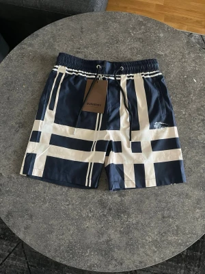 Burberry Shorts Navy Blue - Snygga svorts från Burberry i marinblått med vita grafiska ränder. Klassisk modell med resår och dragsko i midjan. Perfekta för stranden eller poolen, tillverkade i snabbtorkande material. Stilsäker design med Burberry-logga på ena benet.