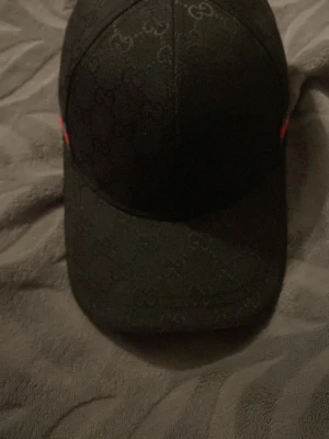 Svart keps från Gucci med GG-mönster - Snygg svart keps från Gucci med diskret GG-monogram över hela kepsen och klassiska röd-gröna ränder på sidan. Böjd skärm och justerbar passform. Perfekt accessoar för en trendig streetstyle-look.
